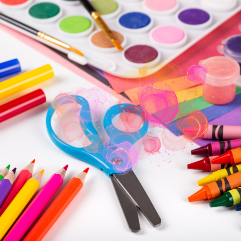 An colorful array of craft supplies including colored pencils, crayons, watercolor paints, markers, paper and a scissors with light blue handles.