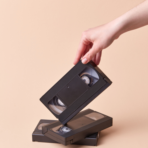 White hand holding VHS tape with two other VHS tapes underneath with tan background.