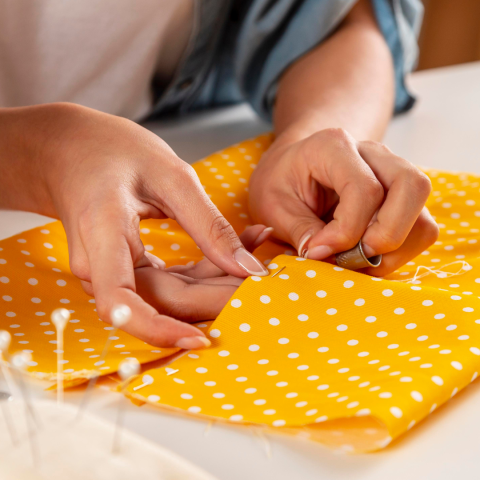 White hands hand sewing a yellow polka dot piece of fabric.
