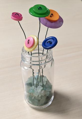 Five button flowers in a small glass jar with green stones on the bottom. Multi-colored buttons are attached to green floral wire. 