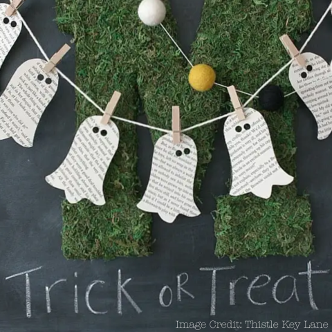 ghost shapes cut out of book pages clipped to a string in front of a blackboard that says "trick or treat" in chalk image credit: thistle key lane