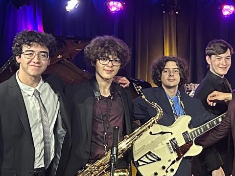 Four young adults stand together smiling, holding instruments, on a stage