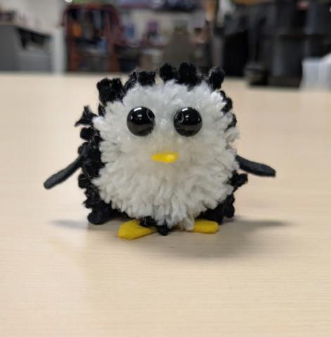 A picture of penguin made out of a pom pom. It has black bead eyes, a yellow felt nose, and yellow felt feet.