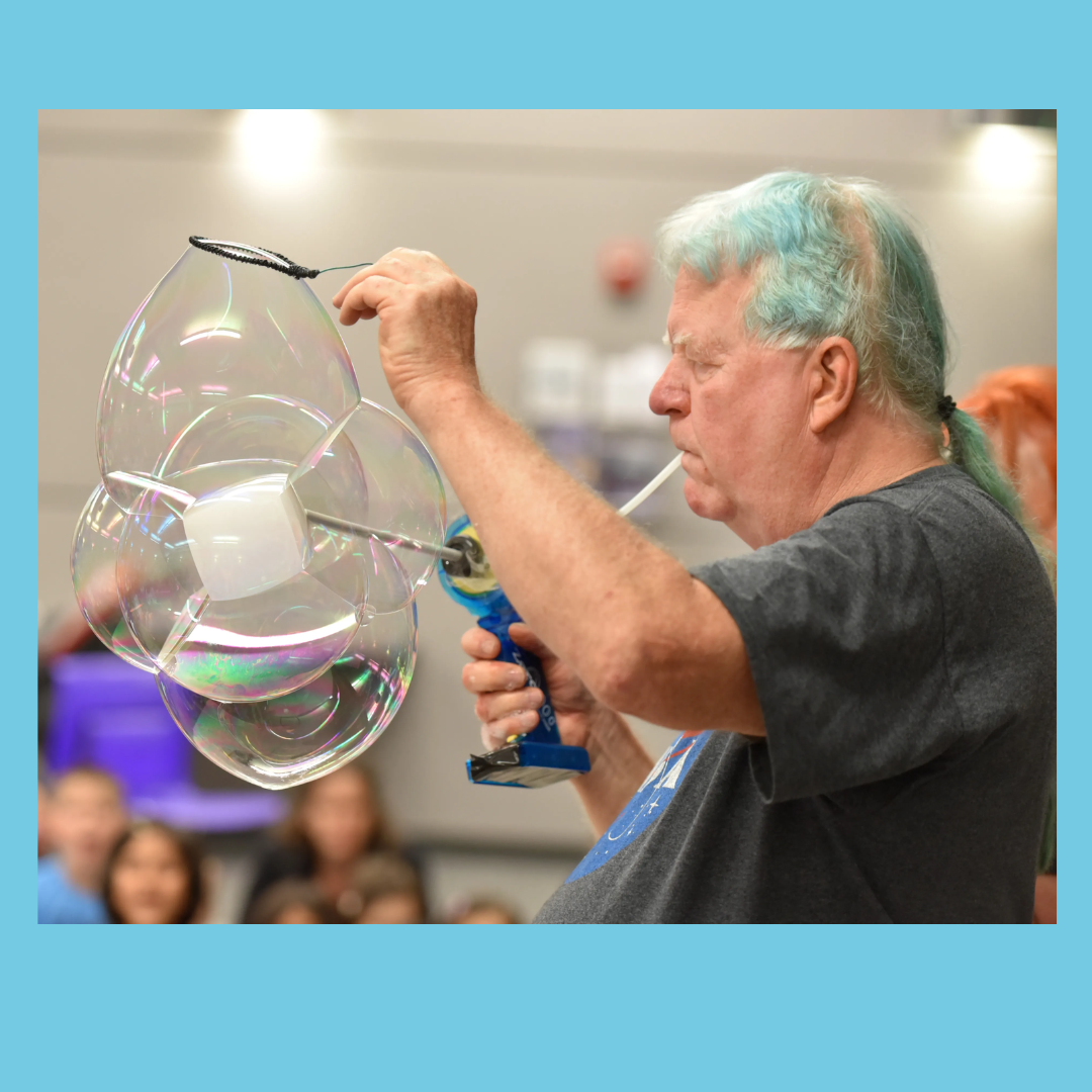 man with blue hair, making large bubbles and using a drill against a blue background